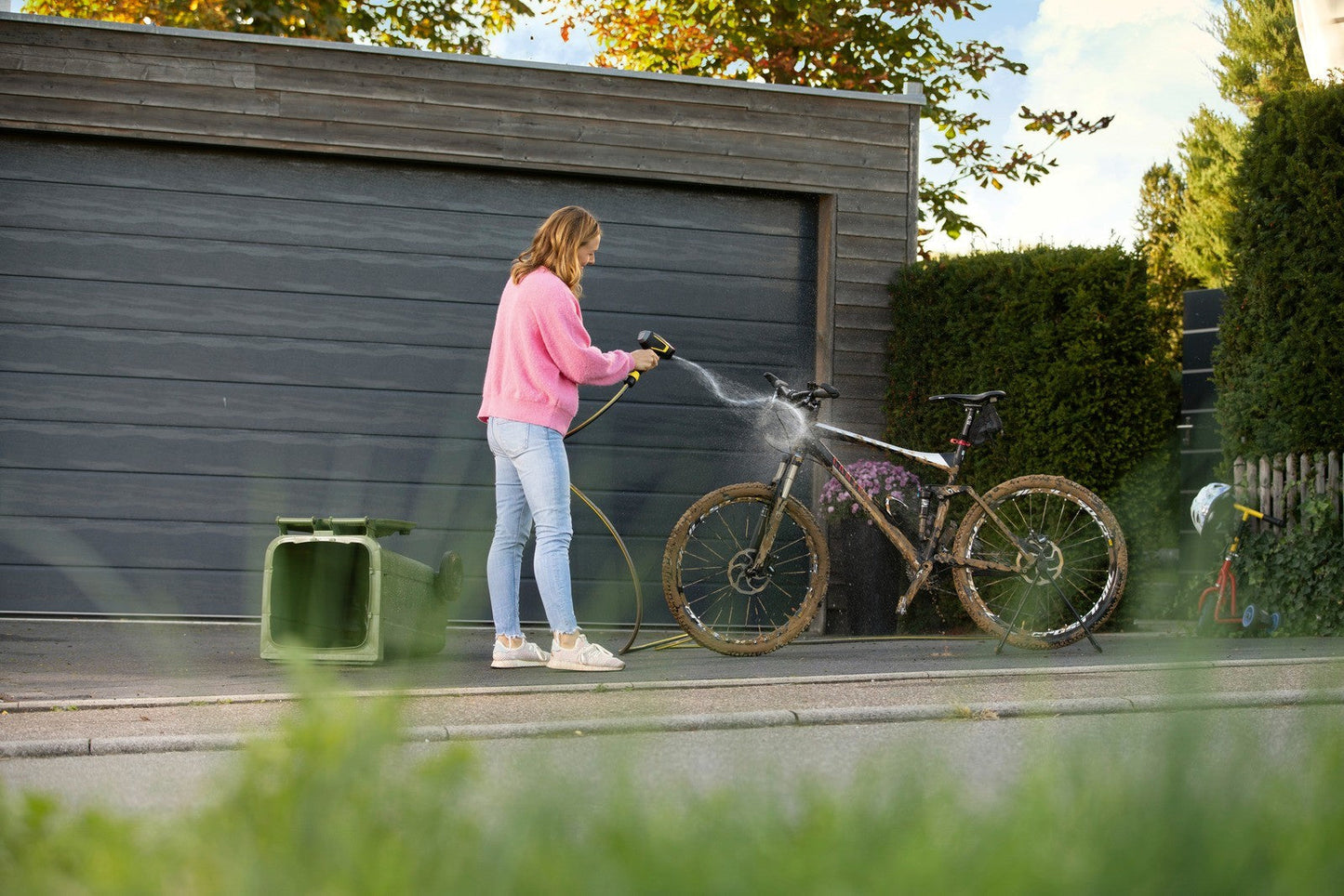 Frau in pinkem Pullover und Jeans mit Kärcher WBS 3 Reinigungspistole auf schmutzigem Mountainbike